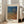 May include: Framed art poster leaning against white wall next to beige sofa in bright living room with large windows. The poster features a dotted, textured pattern in blue and orange tones with Japanese text at top and "YAYOI KUSAMA" at bottom. Light wood frame with white matting, approximately 2-3 feet tall, casting soft shadows on hardwood floor.