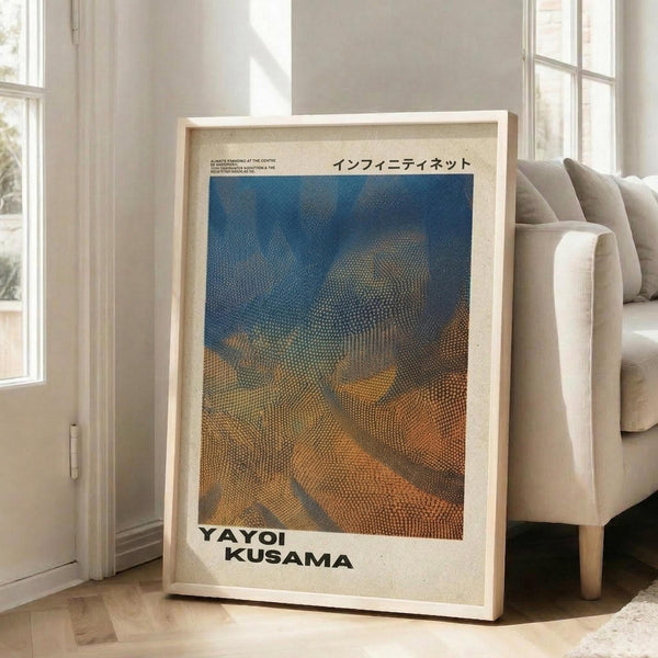 May include: Framed art poster leaning against white wall next to beige sofa in bright living room with large windows. The poster features a dotted, textured pattern in blue and orange tones with Japanese text at top and "YAYOI KUSAMA" at bottom. Light wood frame with white matting, approximately 2-3 feet tall, casting soft shadows on hardwood floor.