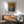 May include: Framed Yayoi Kusama exhibition poster featuring abstract dotted patterns in golden yellow, deep blue, and gray tones displayed above a mid-century modern wooden credenza. Minimalist room setting with mint green upholstered armchair, small round wooden coffee table, white ceramic table lamp, and natural sunlight streaming across white walls.