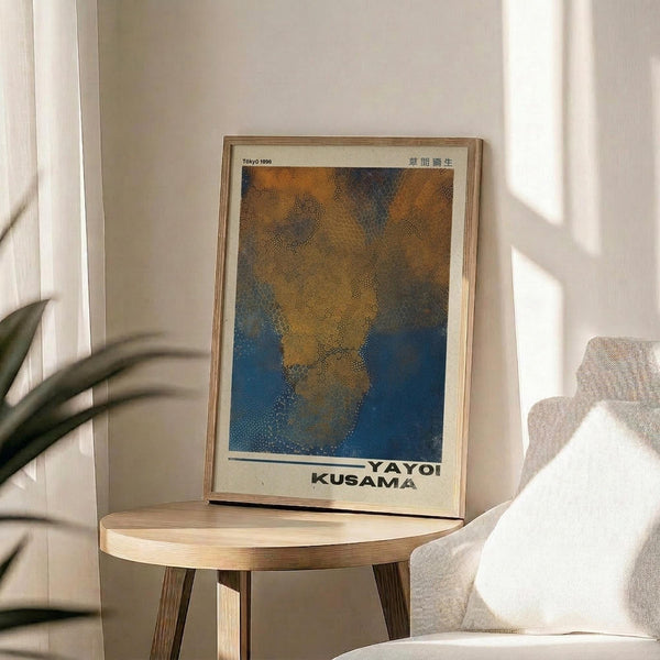 May include: Framed art print displayed on round wooden side table next to beige upholstered chair. The poster features abstract dotted pattern in orange, brown, and blue tones with artist name at bottom. Natural sunlight streams across white walls creating soft shadows. Green plant leaves visible in corner of minimalist room setting.