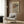 May include: Framed poster with flowing wavy lines in earth tones of rust, olive green, and cream displayed on wood paneled wall above beige upholstered armchair with geometric patterned throw pillow, brass floor lamp, and dark round side table with open book in modern neutral-toned living room setting.