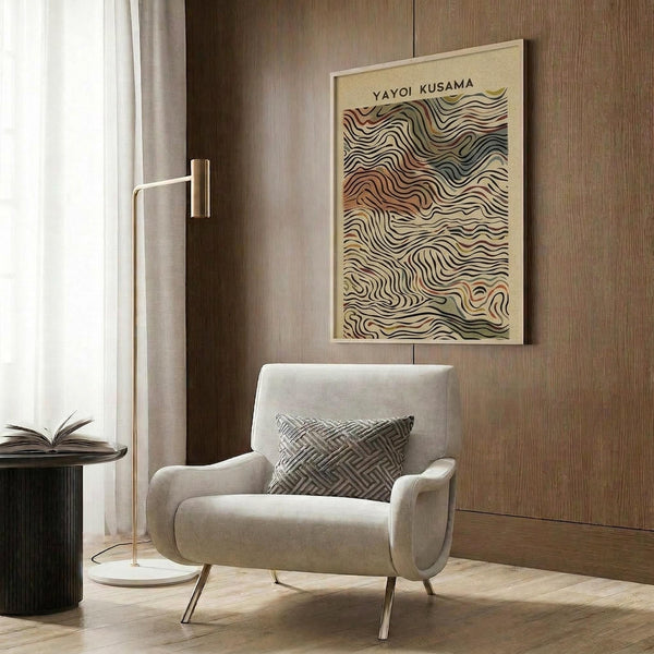 May include: Framed poster with flowing wavy lines in earth tones of rust, olive green, and cream displayed on wood paneled wall above beige upholstered armchair with geometric patterned throw pillow, brass floor lamp, and dark round side table with open book in modern neutral-toned living room setting.