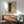 May include: Framed vintage-style poster with black and cream halftone dot pattern creating an abstract curved form, displayed above a wooden sideboard with white ceramic lamp. Mid-century modern living room setting features sage green upholstered armchair with wooden frame, round coffee table, and natural sunlight casting shadows on white wall.