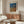 May include: Modern living room with wooden sideboard displaying a framed art print featuring blue and orange dotted landscape patterns with Japanese text. White curved chair, glass coffee table, and brass accent lighting complete the minimalist interior styling.