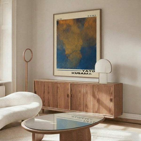 May include: Modern living room with cream walls showcasing a framed poster featuring abstract blue and orange dotted patterns with Japanese text. The artwork hangs above a wooden sideboard with sliding doors, accompanied by a white curved chair, brass floor lamp, round glass coffee table, and minimalist white table lamp.