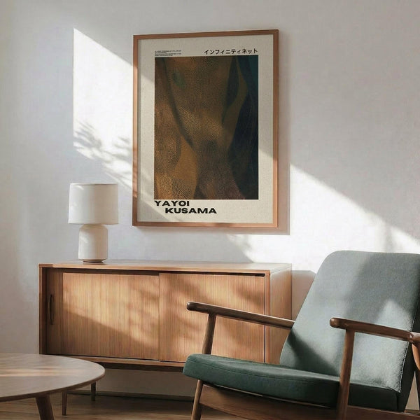 May include: Framed poster print on white wall above wooden sideboard with table lamp. Shows abstract dotted pattern artwork in brown and green tones with Japanese text and "YAYOI KUSAMA" at bottom. Modern living room setting with mint green armchair, round wooden coffee table, and natural sunlight casting shadows across the scene.