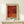 May include: Framed art print with warm red and orange tones featuring abstract dotted patterns and organic shapes, displayed on light wood console table beside small white wrapped gift with greenery sprig, against cream wall with natural lighting