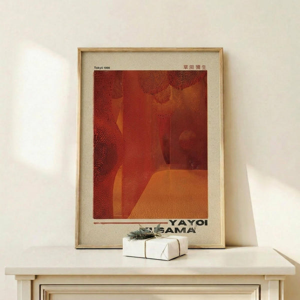 May include: Framed art print with warm red and orange tones featuring abstract dotted patterns and organic shapes, displayed on light wood console table beside small white wrapped gift with greenery sprig, against cream wall with natural lighting