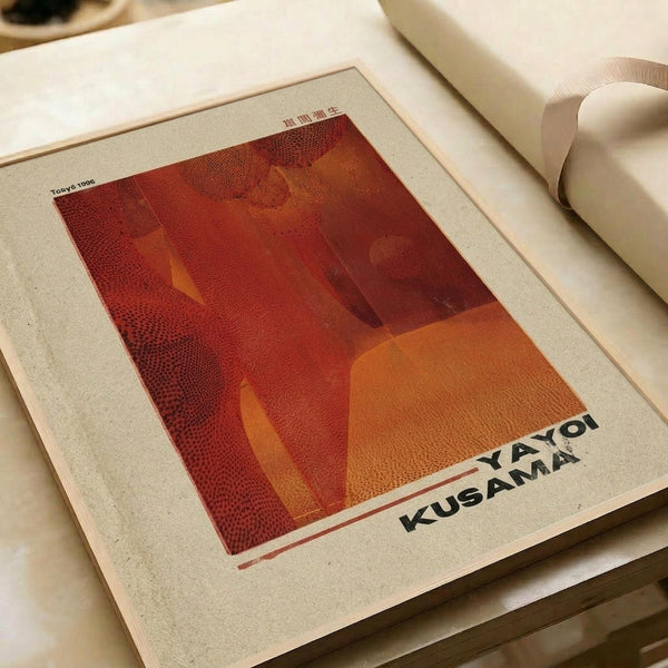 May include: Abstract art print with warm red and orange tones displayed on white paper, featuring geometric shapes and dotted patterns. The artwork shows a stylized figure silhouette against layered colored backgrounds. Text reading "YAYOI KUSAMA" appears at bottom right. Print sits on neutral fabric surface next to white cylindrical objects, creating a minimalist styled scene.