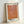 May include: Framed art print leaning against white wall featuring abstract orange and coral gradients with dotted halftone pattern. Japanese text at top, artist name at bottom in black letters. Natural wood frame with white matting, positioned on light surface with soft shadows.
