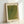 May include: Framed art print leaning against white wall showing green textured abstract pattern with small repeating dots creating flowing organic shapes. Light wooden frame surrounds cream-colored paper with black text at top and Japanese characters on right side. Natural lighting creates soft shadows on wall behind the rectangular vertical poster.