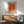 May include: Orange and yellow abstract art poster with Japanese text in wooden frame, displayed above mid-century modern wood credenza with white ceramic lamp. Mint green upholstered chair with wooden arms sits nearby on wooden floor. Warm sunlight creates geometric shadows on white wall in minimalist living room setting.