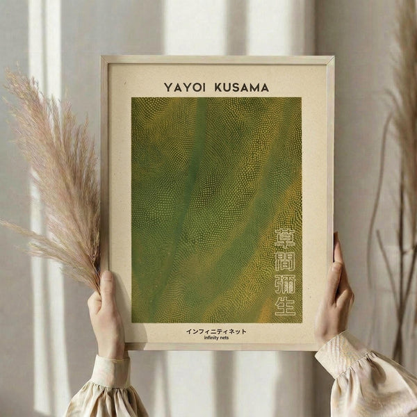 May include: Hands holding framed art print featuring green and yellow dotted pattern in Yayoi Kusama's infinity nets style, with Japanese text below the artwork. Light wooden frame with cream matting, held against neutral beige curtains with dried pampas grass visible on the left side.
