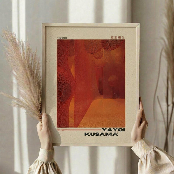 May include: Person holding framed art print with warm orange and red abstract design featuring dotted patterns and flowing forms. Print shows "Tokyo 1998" and "YAYOI KUSAMA" text on cream-colored background. Natural wood frame, held against white curtained window with dried pampas grass visible to the side. Person wearing cream ruffled sleeves.