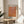 May include: Framed art print with warm orange and coral abstract dotted pattern in thin wooden frame, displayed on round wooden side table next to beige upholstered chair. Soft natural lighting creates shadows on neutral wall background, with plant leaves visible in corner of modern minimalist room setting.