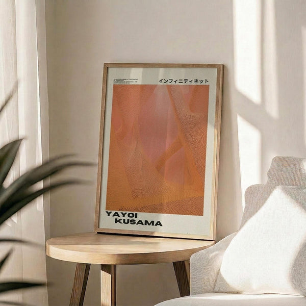 May include: Framed art print with warm orange and coral abstract dotted pattern in thin wooden frame, displayed on round wooden side table next to beige upholstered chair. Soft natural lighting creates shadows on neutral wall background, with plant leaves visible in corner of modern minimalist room setting.