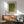 May include: Modern living room setting with sage green upholstered chair, wooden sideboard, and round coffee table. Framed poster on white wall features textured green and yellow pattern with "YAYOI KUSAMA" text and Japanese characters. Natural sunlight casts shadows across the minimalist mid-century furniture arrangement.