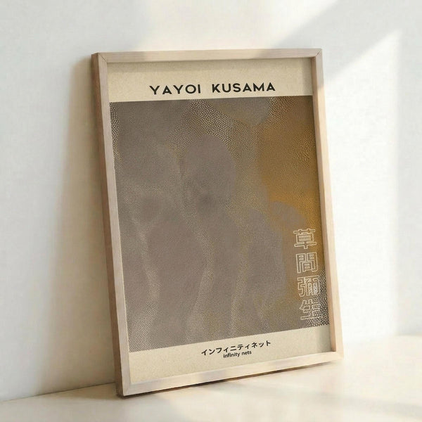 May include: Framed art print leaning against white wall showing abstract dotted pattern in muted browns and grays with Japanese text. Light wood frame with cream matting, captured in soft natural lighting that creates gentle shadows on the wall.