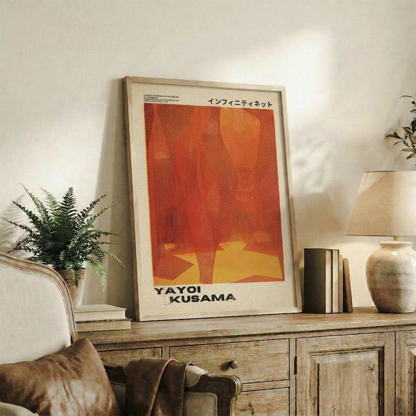May include: Orange and red dotted abstract art poster in light wood frame, displayed on rustic wooden sideboard alongside fern plant, stacked books, and ceramic vases in neutral-toned living room with beige armchair and warm natural lighting.