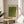 May include: Framed art print displayed on round wooden side table next to beige upholstered chair. Print features green textured pattern with dotted surface resembling reptile skin, Japanese characters in bottom right corner, and "YAYOI KUSAMA" text at top. Natural wood frame matches table. Soft natural lighting creates shadows on white wall, with plant leaves visible in corner.