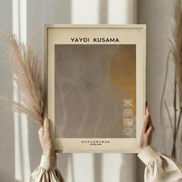 May include: Person in cream sweater holding framed art print with light wood frame, featuring abstract dotted pattern in brown and tan tones with Japanese text. Dried pampas grass visible to left, soft natural lighting against neutral curtained background.