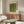 May include: Green textured abstract poster with "YAYOI KUSAMA" text hanging above wooden sideboard in modern living room setting. White curved chair and oval glass coffee table with colorful artwork visible. Warm wood furniture against light walls with natural lighting.