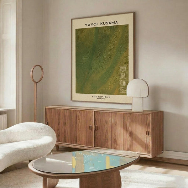 May include: Green textured abstract poster with "YAYOI KUSAMA" text hanging above wooden sideboard in modern living room setting. White curved chair and oval glass coffee table with colorful artwork visible. Warm wood furniture against light walls with natural lighting.