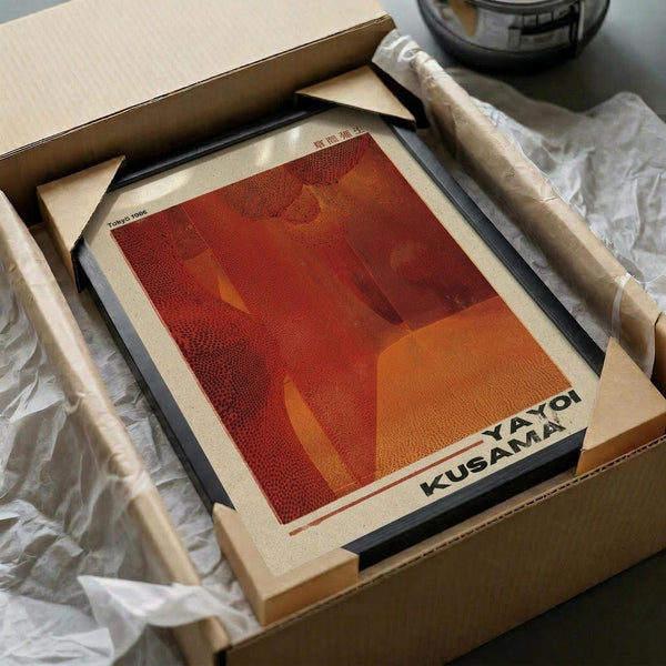 May include: Red and orange abstract art print with geometric shapes and dotted patterns, displayed in black frame on wooden surface. Print shows "KUSAMA YAYOI" text at bottom. Scene includes wooden dowels, white tissue paper, cardboard boxes, and glass container in background, suggesting unboxing or packaging setup.