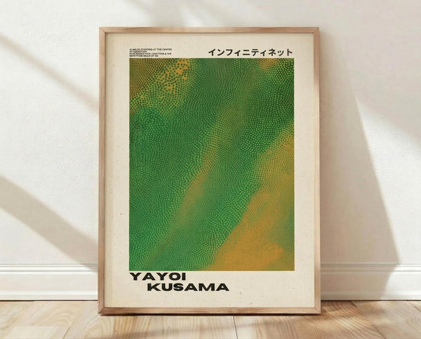 May include: Framed art print leaning against white wall on wooden floor. Light wood frame contains poster with green and yellow dotted pattern resembling organic cellular texture. Japanese text at top, "YAYOI KUSAMA" in bold black letters at bottom. Cream-colored background surrounds the artwork. Natural lighting creates soft shadows on textured white wall behind frame.