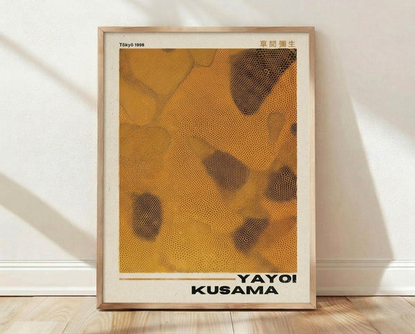 May include: Framed art print leaning against white wall on light wood floor. The poster features abstract orange and yellow dotted patterns with darker spotted areas creating organic, flowing shapes. Text reads "Tokyo 1998" and "YAYOI KUSAMA" in black letters. Natural wood frame with white matting. Soft natural lighting creates gentle shadows on the wall behind the artwork.