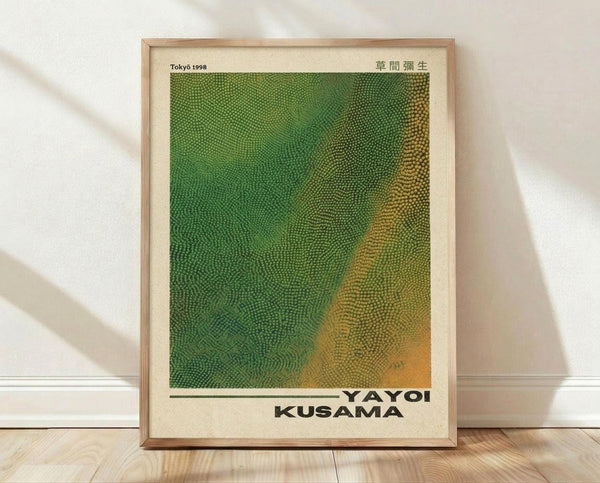 May include: Framed art print with green and yellow dotted pattern creating wavy, flowing abstract design. Features "Tokyo 1998" and "YAYOI KUSAMA" text at bottom with Japanese characters. Light wood frame with cream matting, leaning against white wall on light wooden floor with soft natural lighting casting shadows.