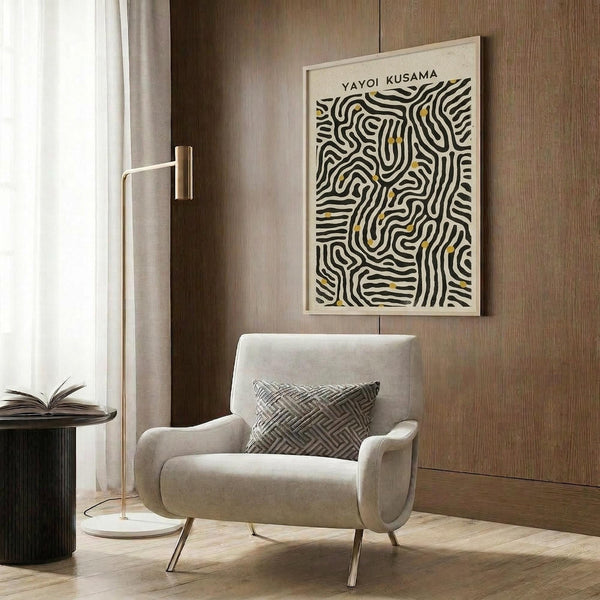 May include: Framed abstract art print with black wavy lines and yellow accents on cream background, displayed on wood paneled wall above beige upholstered chair with geometric patterned pillow. Modern reading corner features brass floor lamp and dark round side table with open book. Warm neutral tones throughout minimalist interior space.