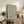 May include: Framed poster with brown polka dot pattern creating optical illusion effect and Japanese text, displayed on rustic wooden sideboard next to fern plant, books, and ceramic vases in warm neutral living room setting with beige armchair visible.