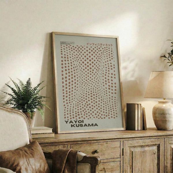 May include: Framed poster with brown polka dot pattern creating optical illusion effect and Japanese text, displayed on rustic wooden sideboard next to fern plant, books, and ceramic vases in warm neutral living room setting with beige armchair visible.