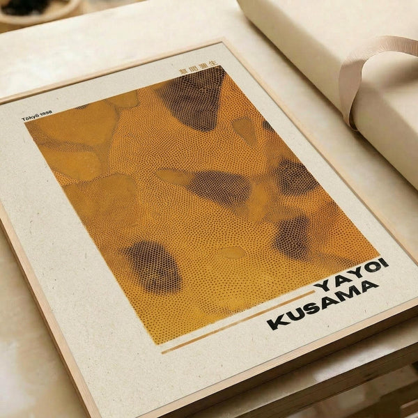 May include: A framed art print featuring a halftone dot pattern artwork in warm orange and brown tones, displayed on white paper with black text reading "YAYOI KUSAMA" at the bottom. The print sits on a beige surface next to rolled white papers, showing how the poster might look when framed and styled in a minimalist workspace setting.
