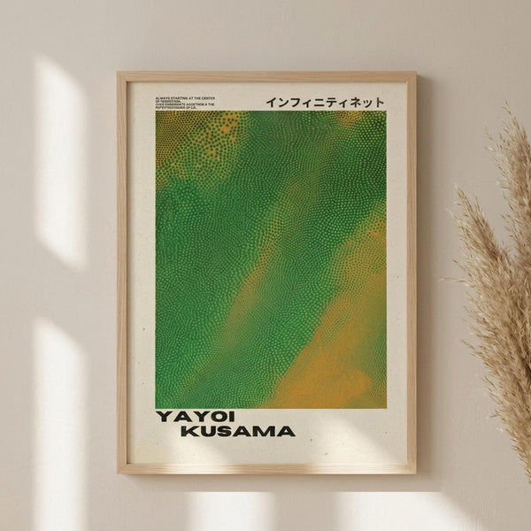 May include: Framed poster with green and yellow dotted pattern resembling Kusama's infinity nets, displayed on beige wall next to dried pampas grass. Light wood frame surrounds white-bordered print featuring Japanese text at top and "YAYOI KUSAMA" at bottom. Natural sunlight casts geometric shadows across the minimal interior setting.