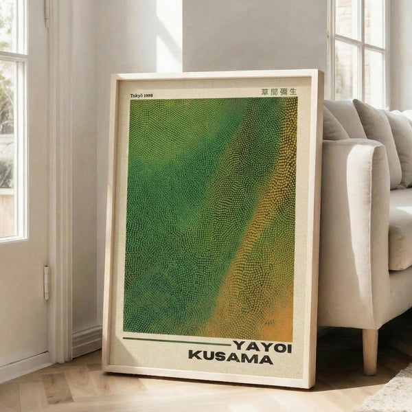 May include: Framed art print leaning against white wall beside beige sofa in bright living room with large windows. The poster features a textured dotted pattern in green, yellow and orange gradient colors with "YAYOI KUSAMA" text at bottom and Japanese characters at top. Light wooden frame, approximately 2-3 feet tall, creates shadow on hardwood floor.