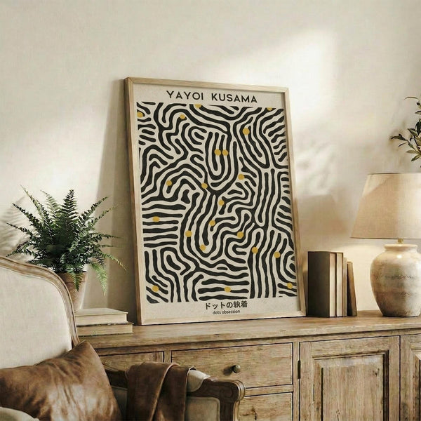 May include: Framed art print with black wavy lines and yellow dots in organic flowing pattern, displayed on rustic wooden sideboard next to fern plant, books, and ceramic vases in warm neutral living room setting with beige armchair visible
