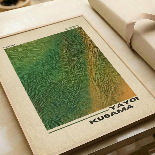 May include: Contemporary art poster print displayed on white surface next to rolled paper tube. Features abstract dotted pattern in green and orange halftone style with "YAYOI KUSAMA" text at bottom. Print has white border and appears to reference 1998 Tokyo exhibition. Warm lighting creates soft shadows on cream-colored background.