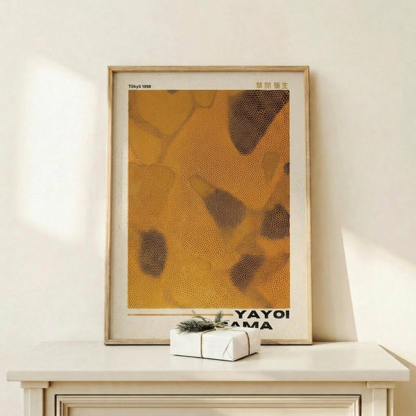 May include: Framed art print in light wood frame leaning against white wall on pale wooden surface. Orange and brown abstract dotted pattern artwork with black text at bottom. Small white wrapped gift box with evergreen sprig sits in front of frame. Minimalist neutral decor setting with natural lighting casting soft shadows.