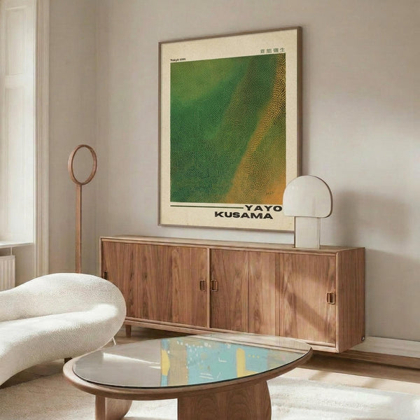 May include: A framed poster with green and yellow textured pattern and "YAYOI KUSAMA" text displayed above a wooden sideboard in a modern living room. The minimalist space features a white sculptural chair, floor lamp, table lamp, and oval coffee table with colorful glass top. Warm natural light creates shadows on pale walls.