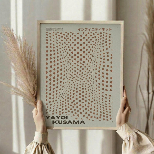 May include: Hands holding a framed poster with brown polka dots creating an optical illusion pattern on cream background. Japanese text appears at top. Person wears beige ruffled sleeves and holds dried wheat stems. Natural lighting creates soft shadows on neutral wall.