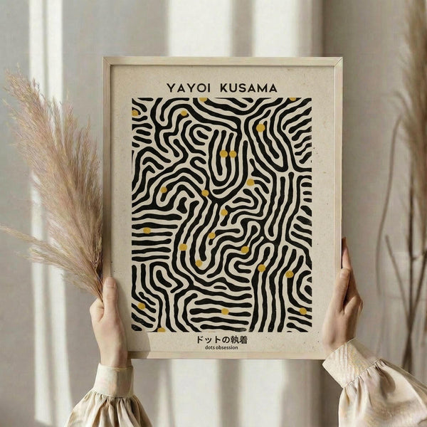 May include: Person holding a framed art print with black wavy maze-like lines and small yellow dots on cream paper. Light wood frame, neutral background with dried pampas grass visible. Hands wearing beige ruffled sleeves grip the rectangular poster displaying abstract flowing patterns reminiscent of topographic maps or organic cellular structures.