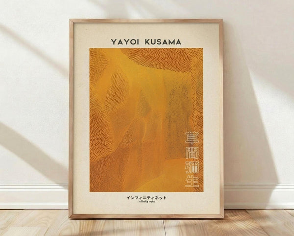 May include: Framed art print leaning against white wall on light wood floor. Warm orange artwork features flowing dotted patterns reminiscent of cellular structures or organic networks. Black text at top reads "YAYOI KUSAMA" with Japanese characters vertically aligned on right side and "infinity nets" at bottom. Natural wood frame surrounds cream-colored matting. Soft natural lighting creates gentle shadows on textured white wall.