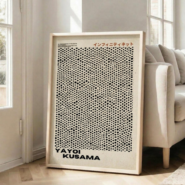 May include: Framed art print with black polka dot pattern on cream background, featuring Japanese text and "YAYOI KUSAMA" at bottom, displayed in light wood frame leaning against white wall next to beige upholstered chair in bright living room with large windows