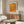 May include: Orange and yellow dotted abstract art poster with Japanese text hanging above a wooden sideboard in a minimalist living room. White curved chair and glass coffee table visible in foreground. Warm wood tones throughout the mid-century modern styled space with white lamp and natural lighting.