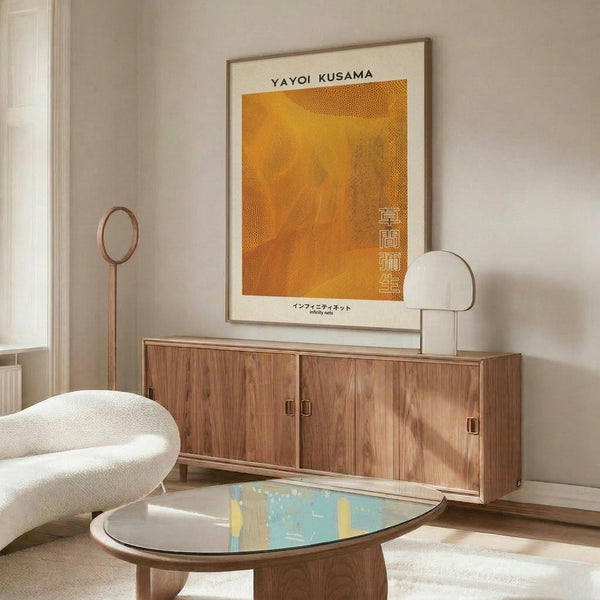 May include: Orange and yellow dotted abstract art poster with Japanese text hanging above a wooden sideboard in a minimalist living room. White curved chair and glass coffee table visible in foreground. Warm wood tones throughout the mid-century modern styled space with white lamp and natural lighting.