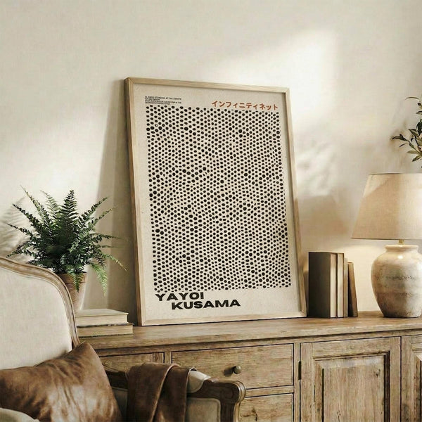 May include: Framed poster with black polka dot pattern on white background displayed on wooden sideboard next to fern plant, books, and ceramic vases in neutral-toned living room setting with beige armchair visible in foreground