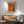 May include: A framed art poster with orange, brown and green dotted abstract design hanging above a wooden sideboard. Mid-century modern living room setting with gray upholstered armchair, small white table lamp, and round coffee table. Natural sunlight creates shadows across white walls. Warm wood furniture complements the earthy tones in the artwork.