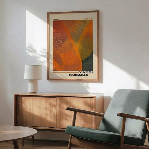 May include: A framed art poster with orange, brown and green dotted abstract design hanging above a wooden sideboard. Mid-century modern living room setting with gray upholstered armchair, small white table lamp, and round coffee table. Natural sunlight creates shadows across white walls. Warm wood furniture complements the earthy tones in the artwork.